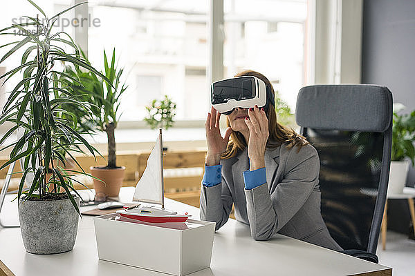 Geschäftsfrau sitzt am Schreibtisch mit einem Schiffsmodell und schaut durch eine VR-Brille