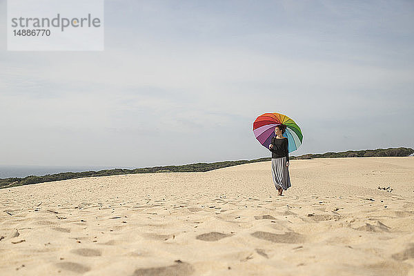 Frau mit buntem Regenschirm steht auf einer Düne und schaut in die Ferne