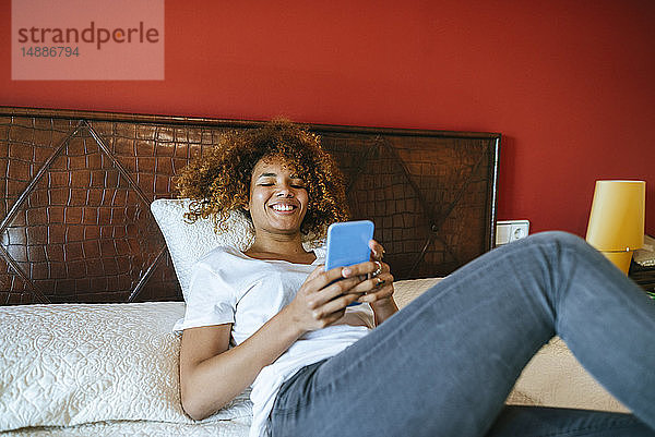 Glückliche junge Frau mit lockigem Haar liegt zu Hause im Bett und benutzt ihr Handy