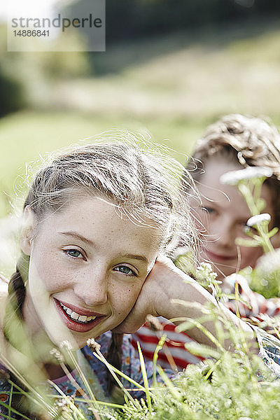 Porträt eines lächelnden Mädchens auf einer Wiese liegend mit kleinem Bruder im Hintergrund