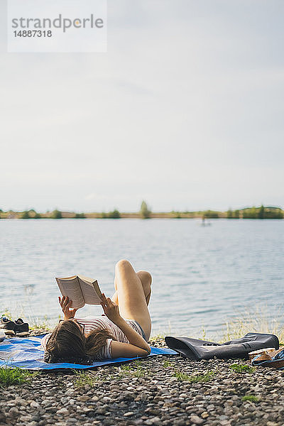 Junge Frau liegt auf einer Decke am Seeufer und liest ein Buch