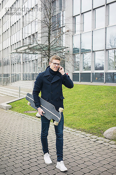 Lächelnder junger Mann mit Skateboard beim Telefonieren mit dem Handy in der Stadt