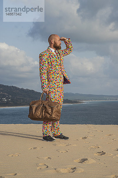 Spanien  Tarifa  Mann mit Reisetasche im Anzug mit bunten Polka-Punkten steht auf einer Düne und schaut in die Ferne