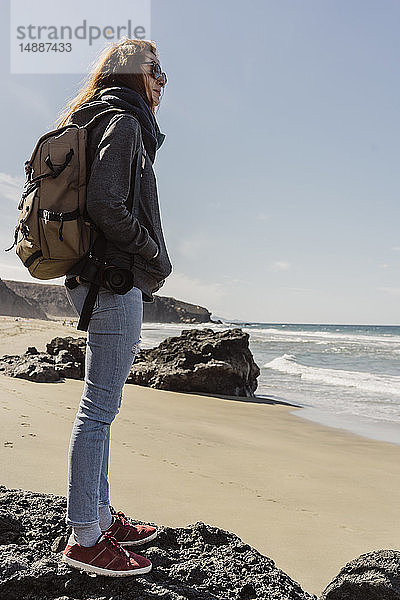 Spanien  Kanarische Inseln  Fuerteventura  junge Frau mit Rucksack steht am Strand und schaut in die Ferne