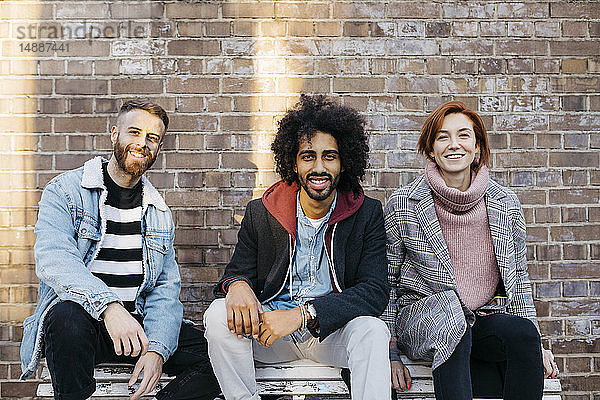 Porträt von drei glücklichen Freunden  die auf einer Bank vor einer Ziegelmauer sitzen