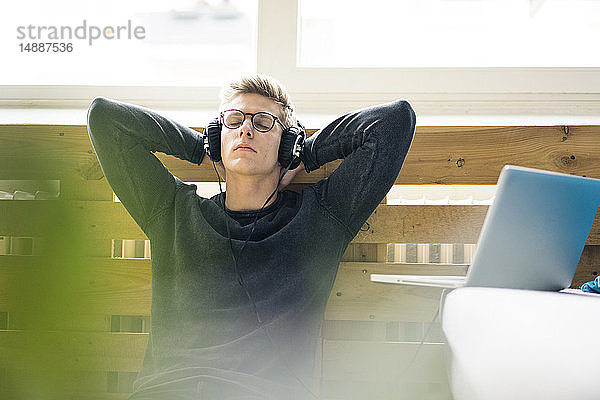Entspannter junger Mann sitzt auf dem Boden und hört Musik mit Kopfhörern