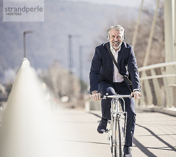 Lächelnder reifer Geschäftsmann mit Kopfhörern beim Fahrradfahren auf einer Brücke in der Stadt