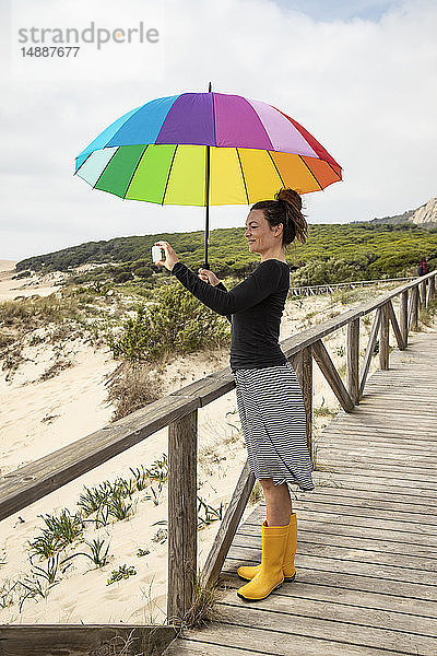 Frau mit buntem Regenschirm steht am Strand und nimmt einen Selfie