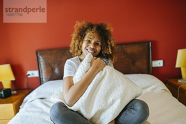 Porträt einer glücklichen jungen Frau mit lockigem Haar  die zu Hause auf dem Bett sitzt und ein Kissen hält