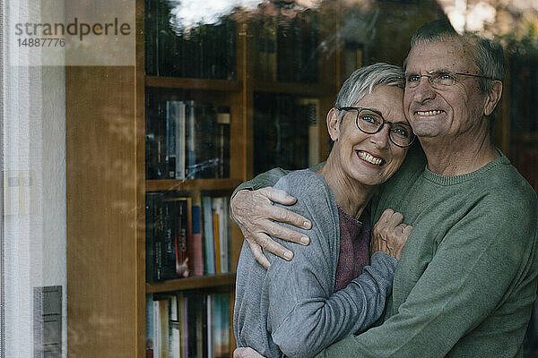 Porträt eines glücklichen älteren Paares  das sich hinter einer Fensterscheibe umarmt