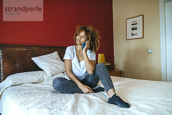 Junge Frau mit lockigem Haar sitzt im Bett und telefoniert mit dem Handy