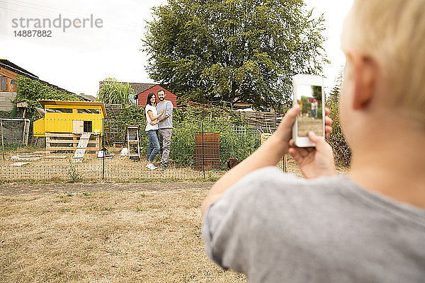 Junge macht Handyfoto von Eltern  die im Hühnerstall im Garten stehen