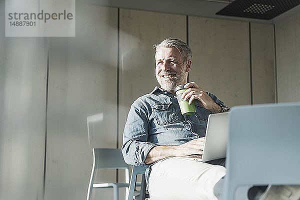 Lächelnder  lässiger  reifer Geschäftsmann mit Laptop und Kaffee zum Mitnehmen