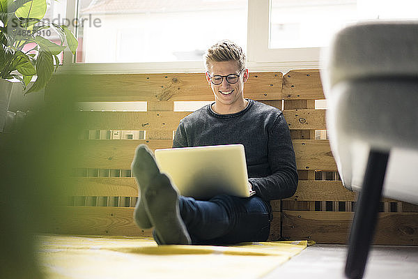 Lächelnder junger Mann sitzt mit Laptop auf dem Boden