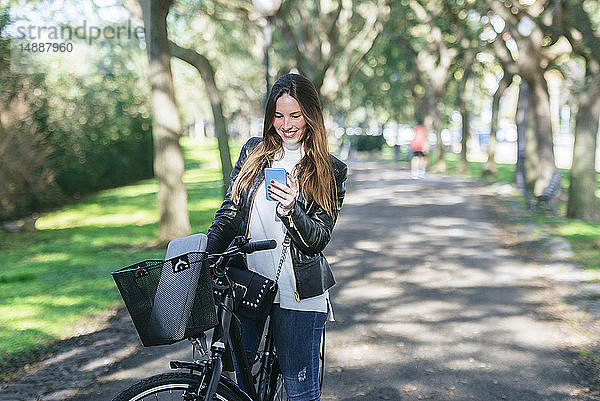 Lächelnde junge Frau mit Fahrrad im Park mit Mobiltelefon