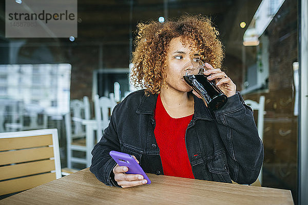 Junge Frau mit Handy trinkt Cola in einer Bar