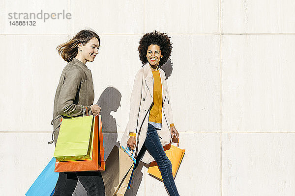 Zwei glückliche Frauen mit Einkaufstaschen  die an einer Wand entlanggehen