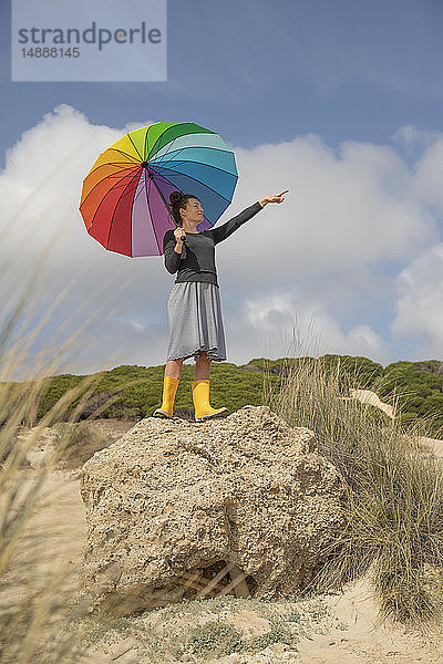 Frau mit buntem Regenschirm steht auf einem Felsen und schaut in die Ferne