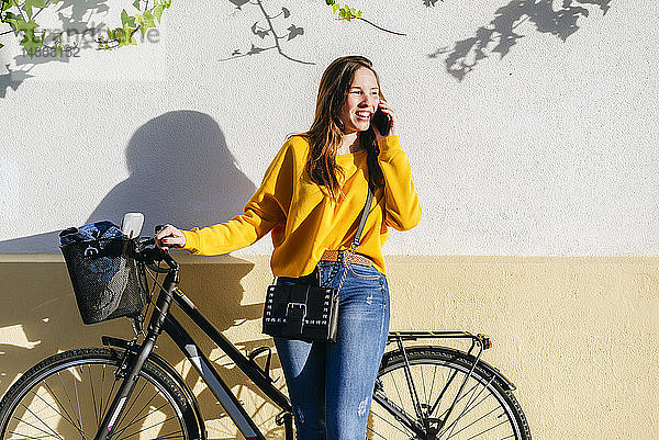 Junge Frau mit Fahrrad telefoniert mit dem Handy an einer Wand