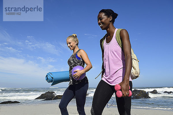 Zwei glückliche Frauen mit Fitnessgeräten am Strand spazieren