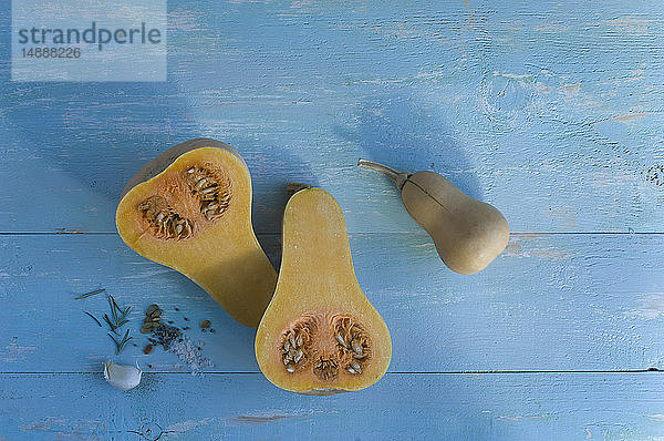 Ganzer und in Scheiben geschnittener Butternusskürbis  Knoblauchzehe und Kräuter auf hellblauem Holz
