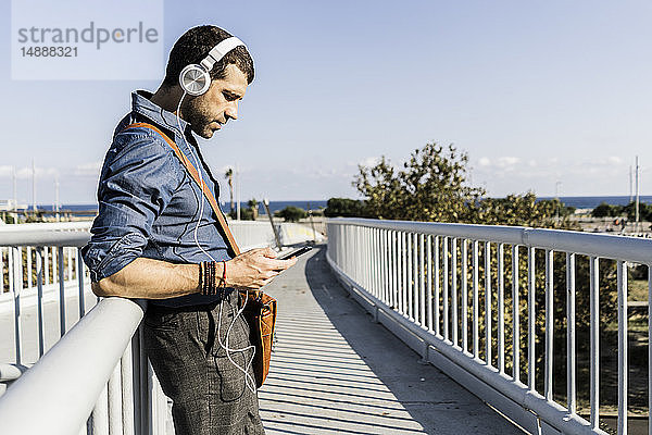 Mann steht auf Steg und hört Musik mit Kopfhörern und Smartphone