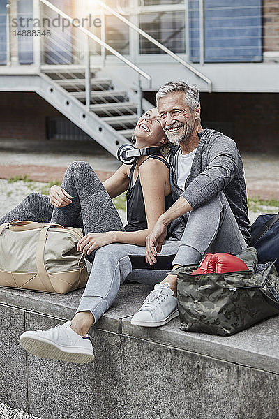 Lachender reifer Mann und junge Frau mit Sporttasche vor der Turnhalle sitzend