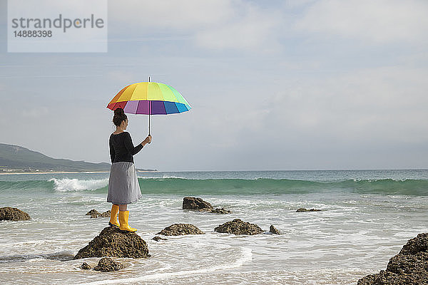 Frau mit buntem Regenschirm auf einem Stein am Strand stehend