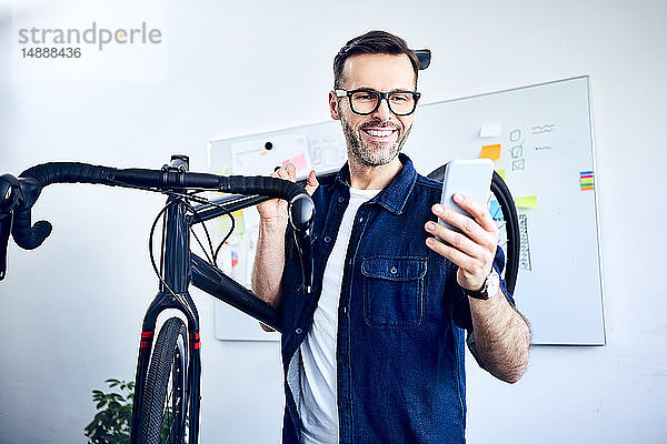 Lächelnder Geschäftsmann trägt Fahrrad im Büro und schaut auf Smartphone