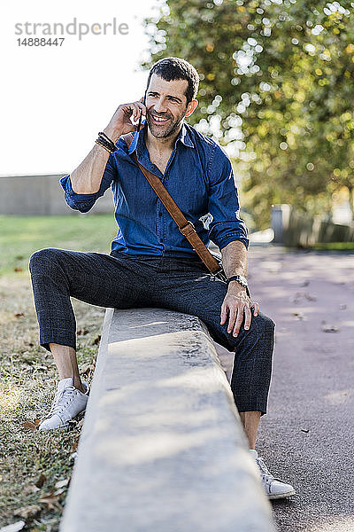 Porträt eines lächelnden Mannes am Telefon  der im Freien an einer Wand sitzt