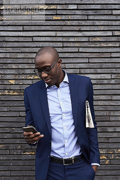 Porträt eines lächelnden Geschäftsmannes mit Zeitung  Brille und blauem Anzug  der auf sein Mobiltelefon schaut