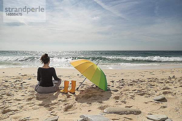 Frau mit buntem Sonnenschirm am Strand sitzend  Rückansicht