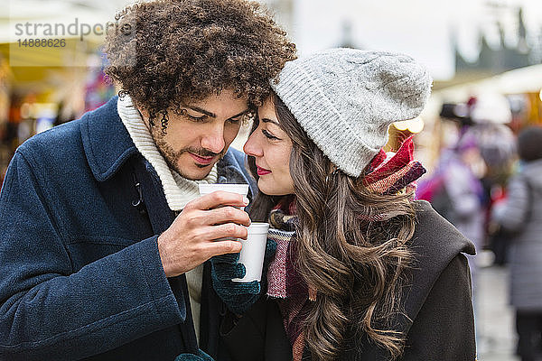 Glückliches  anhängliches junges Paar mit heißen Getränken auf dem Weihnachtsmarkt