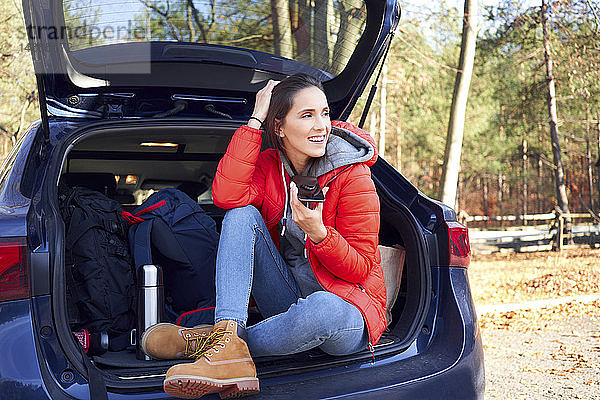 Glückliche Frau  die während einer Autofahrt ein Smartphone benutzt  während sie im Kofferraum sitzt