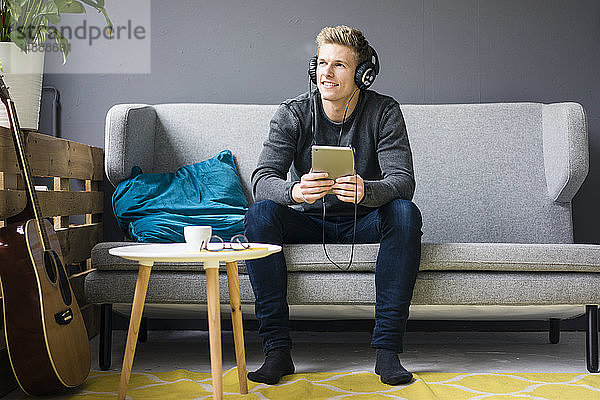 Lächelnder junger Mann mit Gitarre  Tablett und Kopfhörer auf Couch sitzend