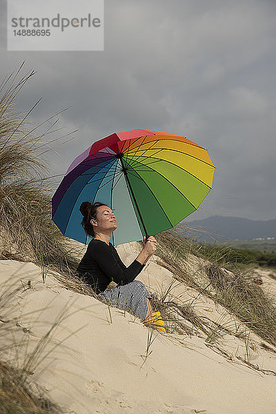 Frau mit buntem Sonnenschirm am Strand sitzend  sonnenbadend