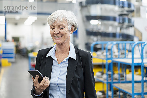 Lächelnde ältere Geschäftsfrau betrachtet Handy in einer Fabrik