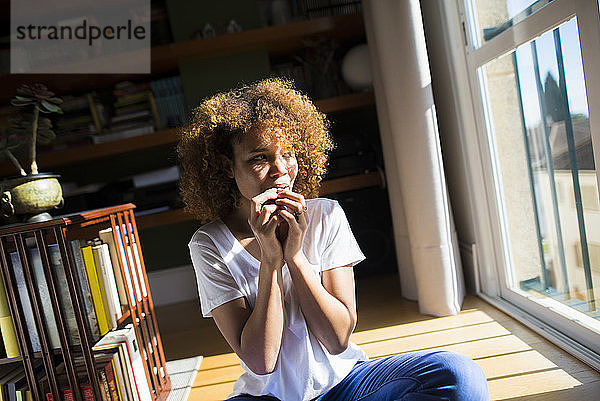 Junge Frau mit lockigem Haar sitzt bei Sonnenschein zu Hause auf dem Boden