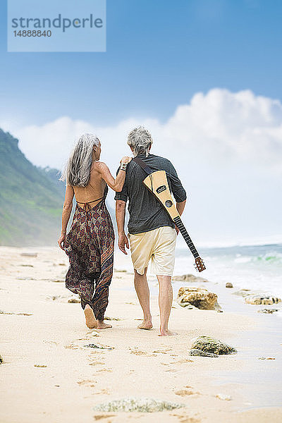 Rückenansicht eines älteren Hippie-Paares mit Gitarre  das Seite an Seite am Strand spazieren geht