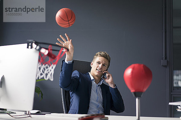 Junger Geschäftsmann sitzt am Schreibtisch im Büro  spielt Basketball und telefoniert