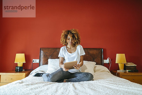 Junge Frau mit lockigem Haar sitzt mit Tablette auf dem Bett