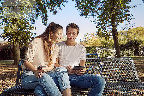 Junges Paar im Park sitzend auf einer Bank mit Handy und Tablett