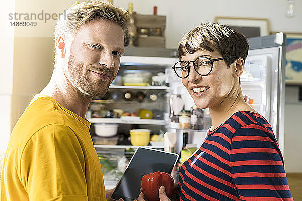 Porträt eines Paares mit Tablette vor dem Kühlschrank beim Online-Einkauf von Lebensmitteln