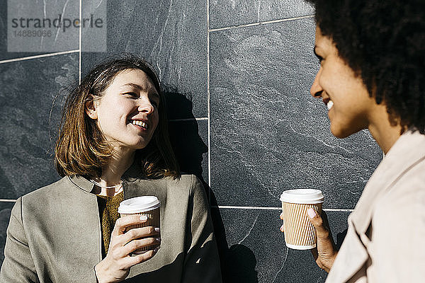 Zwei glückliche Frauen mit Kaffee zum Mitnehmen sprechen gegen eine Wand