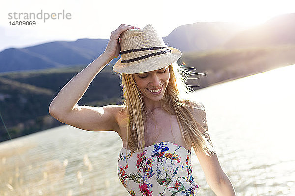 Lächelnde junge Frau mit Sommerhut und Top mit Blumenmuster vor dem See