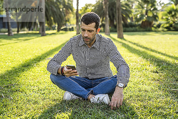 Mann sitzt auf einer Wiese im Stadtpark und schaut auf sein Handy