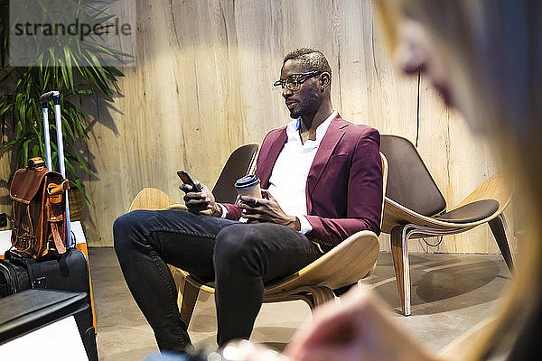 Geschäftsmann mit Gepäck sitzt in der Hotellobby  benutzt ein Smartphone  trinkt Kaffee