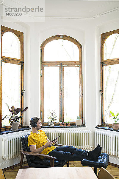 Lässiger Mann in gelbem Hemd mit Kopfhörern sitzt im Lounge Chair in stilvoller Wohnung