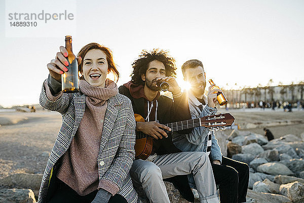Drei glückliche Freunde bei Sonnenuntergang mit Gitarre auf Bierflaschen anstoßen