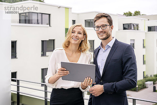 Immobilienmakler steht mit einem Kunden auf einem Balkon und schaut auf ein digitales Tablett